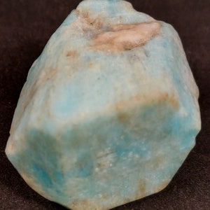 Puede incluir: Un gran cristal de aguamarina azul claro con una superficie rugosa y natural. El cristal tiene una apariencia ligeramente nublada con algunas inclusiones blancas y marrones.