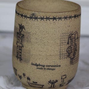 May include: A speckled tan ceramic tumbler featuring black line art illustrations of two jester-like figures, a barbed wire border, and various small household icons. The text "ladybug ceramics made in chicago" is printed in black on the front.