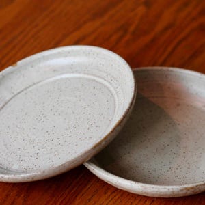May include: Two off-white ceramic plates with speckled detailing. The plates have a shallow, oval shape with a slightly raised rim. The plates are set against a warm, wooden surface, showcasing their neutral tones and rustic aesthetic.