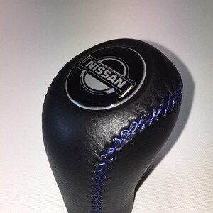 Può includere: Pomello del cambio in pelle nera con cuciture blu e logo Nissan argentato.