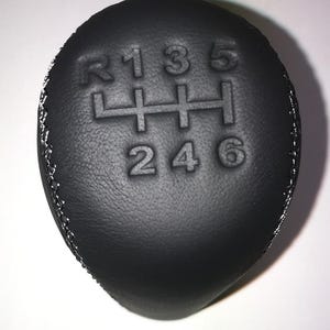 Op de afbeelding: Zwarte lederen versnellingspookknop met witte stiksels. De knop toont het schakelpatroon: R, 1, 3, 5 en 2, 4, 6. De versnellingspookknop is een vervangend onderdeel voor een voertuig.