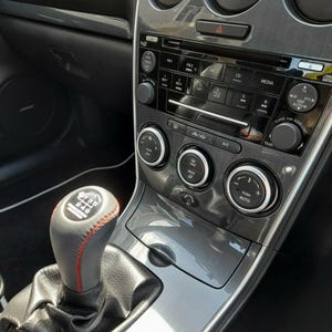 Peut inclure: Intérieur d'une voiture avec tableau de bord et console. Le tableau de bord noir avec des garnitures argentées comprend une radio, des bouches d'aération et des commandes de climatisation. Un levier de vitesse noir avec des coutures rouges est visible.
