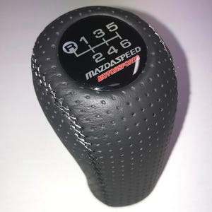 May include: A black leather gear stick knob with white stitching and a perforated surface. The top has a black circular emblem displaying a gear shift pattern and the text "MAZDASPEED MOTORSPORTS".