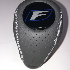 May include: Gray leather gear shift knob with white stitching and a black circle with a silver "F" logo in a blue outline.