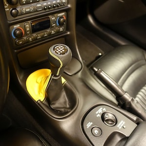 Könnte beinhalten: Ein schwarzer Leder-Schalthebelknauf mit gelben Nähten und einem 6-Gang-Schaltmuster. Der Knauf befindet sich im Innenraum eines Autos mit einem schwarzen Ledersitz und einem schwarzen Armaturenbrett.