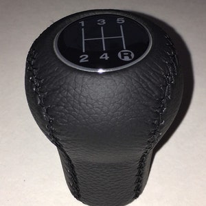 Może przedstawiać: Czarny skórzany gałka zmiany biegów z czarnym i srebrnym wzorem biegów. Wzór biegów pokazuje liczby 1, 2, 3, 4, 5 i literę R.