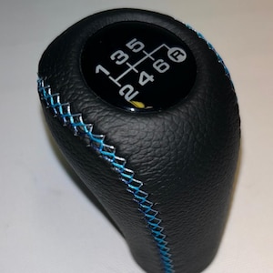 May include: Black leather gear stick knob with blue stitching. The top has a black surface with white numbers indicating gear positions. The gear stick knob is designed for a manual transmission.