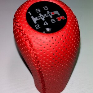 Pode incluir: Botão de câmbio de couro vermelho com logotipo Nismo preto e padrão de 6 velocidades.