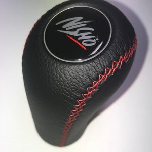 Peut inclure: Pommeau de levier de vitesses en cuir noir avec surpiqûres rouges. Le dessus présente un cercle noir avec du texte blanc et rouge. Le pommeau est pour le changement de vitesse d'un véhicule.