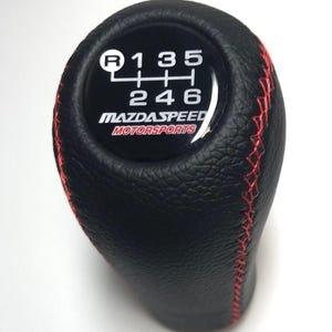 Könnte beinhalten: Schwarzer Lederschaltknauf mit roten Nähten. Die Oberseite zeigt ein Schaltschema und das MazdaSpeed Motorsports-Logo. Der Knauf hat eine strukturierte Oberfläche und ist für ein Fahrzeug mit Schaltgetriebe konzipiert.