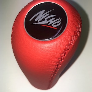 May include: Red leather gear shift knob with a black circular top featuring the word "NISMO" in a stylised silver font.