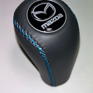 Könnte beinhalten: Schwarzer Schalthebelknauf mit blauen Nähten und Mazda-Logo. Der Knauf besteht aus einem glatten, dunklen Material und verfügt über ein rundes Emblem mit dem Mazda-Logo in Weiß auf schwarzem Hintergrund.