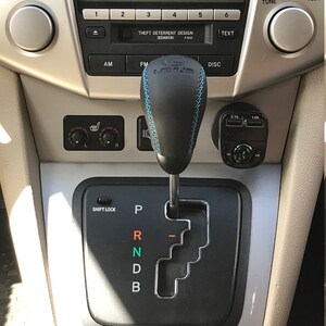 Op de afbeelding: Een zwarte leren versnellingspookknop met blauwe stiksels en een zilveren versnellingspook. De versnellingspook heeft de letters P, R, N, D en B erop. De versnellingspook zit in een auto met een zilveren dashboard.