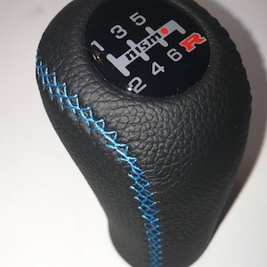 May include: Black leather gear shift knob with blue stitching and a black and silver gear pattern with the word "nismo" and the letter "R" in red.