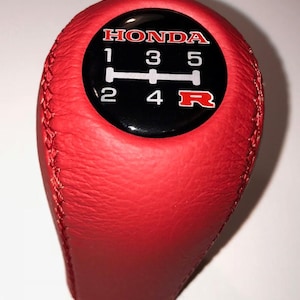 May include: Red leather Honda gear stick knob with a black circular emblem. The emblem shows the Honda logo and the gear shift pattern: 1, 2, 3, 4, 5, and R. Designed for a manual transmission.