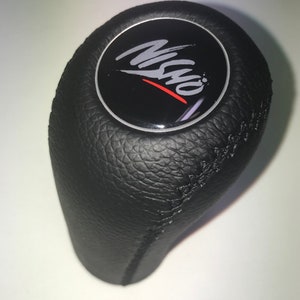 Op de afbeelding: Zwarte leren versnellingspookknop met een zilveren en zwart rond embleem met de tekst "Misho" in een gestileerd lettertype met een rode lijn.
