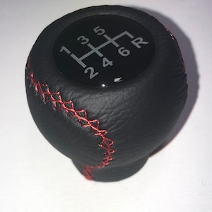 May include: Black gear shift knob with red stitching. The top of the knob has a diagram of a 6-speed manual transmission gear pattern, with numbers 1 through 6 and reverse (R).