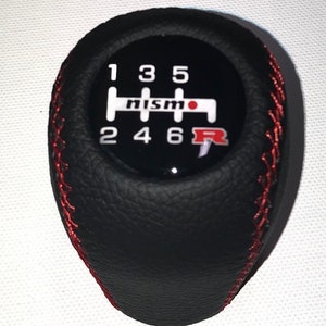 Może przedstawiać: Czarny skórzany gałka zmiany biegów z czerwonymi szwami i czarno-białym logo NISMO z czerwoną kropką. Gałka ma biały wzór biegów z numerami 1, 3, 5, 2, 4, 6 i literą R.
