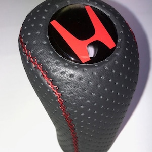 Peut inclure: Pommeau de levier de vitesse en cuir noir perforé avec surpiqûres rouges. Il présente un logo "H" rouge sur une surface noire brillante. Le pommeau est conçu pour le changement de vitesse d'un véhicule.