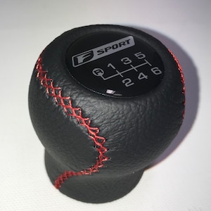 Peut inclure: Pommeau de levier de vitesse en cuir noir avec surpiqûres rouges et un logo F SPORT noir et argent avec un schéma de changement de vitesse.