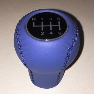 May include: A blue leather gear stick knob with a black top. The top features a diagram of a 6-speed manual transmission gear pattern. The knob is stitched with a contrasting thread.