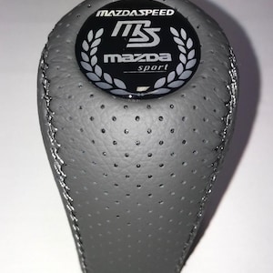 Peut inclure: Pommeau de levier de vitesse MazdaSpeed en cuir gris avec un logo noir et blanc. Le logo comporte le texte "MAZDASPEED" au-dessus d'un emblème "MS" stylisé et "mazda sport" en dessous. Le pommeau a une texture perforée et des coutures argentées.