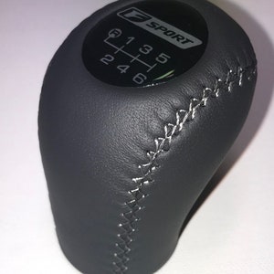 May include: Black leather gear shift knob with a stitched seam and a black F SPORT logo with white lettering and a white gear shift pattern.