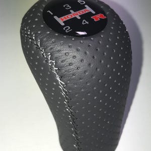 May include: A black perforated leather gear stick knob with white stitching. The top has a black circular insert with the Honda logo and gear shift pattern in white and red. The gear stick knob is designed for a car.