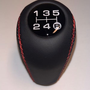 May include: Black leather gear shift knob with red stitching. The top features a black circle with white numbers and a gear shift pattern: 1, 2, 3, 4, 5, and R for reverse.