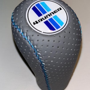 Op de afbeelding: Grijze geperforeerde versnellingspookknop met blauwe stiksels. De knop heeft een cirkelvormig embleem met de tekst "4Runner" en blauwe strepen. De versnellingspookknop is ontworpen voor een voertuig.