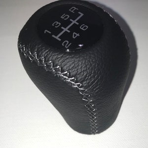 May include: Black leather gear shift knob with white stitching and a black plastic top with white numbers 1, 2, 3, 4, 5, 6, and R.