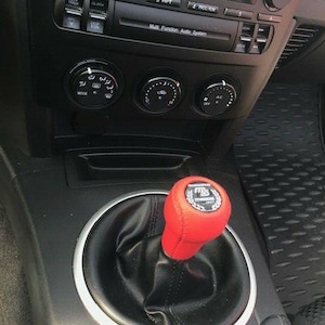 Puede incluir: Un pomo de palanca de cambios de cuero rojo con un emblema plateado, colocado en una bota de cuero negro. El salpicadero incluye un sistema de audio multifunción y controles de climatización.
