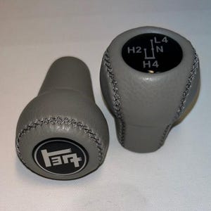 Può includere: Due pomelli del cambio grigi con superfici testurizzate e dettagli a punto croce. Un pomello presenta una parte superiore circolare nera con il testo H2, H4, N e L4. L'altro mostra un emblema circolare nero con un logo grafico bianco stilizzato.