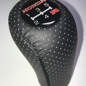 May include: Black perforated leather Honda gear stick knob with white stitching. The top has a black face with white gear numbers and a red "R" for reverse. The word "HONDA" is in red.