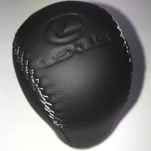 Op de afbeelding: Zwarte leren versnellingspookknop met witte stiksels en het Lexus-logo in reliëf op de bovenkant.