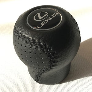 Op de afbeelding: Zwarte leren versnellingspookknop met een zilveren Lexus-logo. De knop heeft een geperforeerd ontwerp.