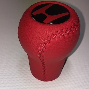 Può includere: Pomello del cambio in pelle rossa con un logo Honda nero e rosso in cima. Il pomello ha una cucitura sul lato.