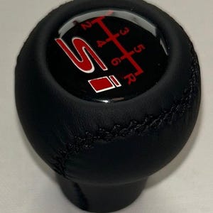 Könnte beinhalten: Schwarzer Lederschaltknauf mit rotem Gangschaltmuster und "SI"-Logo. Das Muster umfasst die Zahlen 1 bis 6 und "R" für den Rückwärtsgang. Der Knauf hat eine Naht und eine glänzende Oberseite.