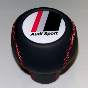 Puede incluir: Pomo de cambio de marchas de cuero negro con costuras rojas y un emblema circular blanco con rayas rojas, blancas y negras y el texto "Audi Sport".