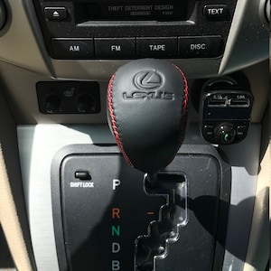 Op de afbeelding: Een zwarte leren versnellingspookknop met rode stiksels en een zilveren Lexus-logo. De versnellingspook heeft de letters P, R, N, D en B.