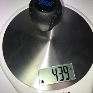 May include: A black leather gear shift knob with a blue "STI" logo on top, weighing 430 grams on a digital scale.