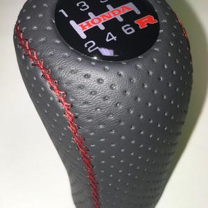 Peut inclure: Un pommeau de levier de vitesse Honda noir et rouge avec une texture en cuir perforé. Le schéma de changement de vitesse est affiché sur une surface circulaire noire, avec le logo Honda en rouge. Le pommeau a des coutures rouges.