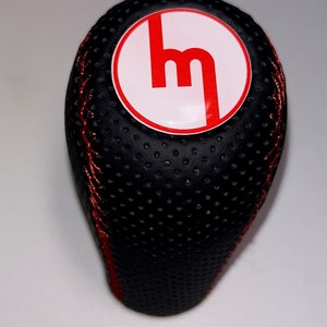 Peut inclure: Pommeau de levier de vitesse en cuir noir avec surpiqûres rouges et un cercle blanc avec un logo "M" rouge.