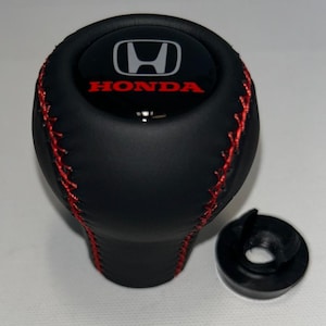 May include: A black gear shift knob featuring red cross-stitching along the sides and a circular top insert displaying the silver Honda logo above the red Honda text. A separate black plastic circular adapter piece sits to the right of the knob.
