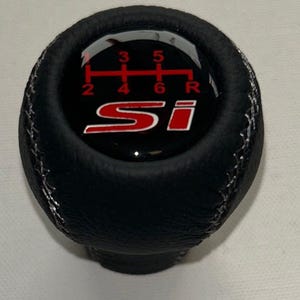 Puede incluir: Pomo de palanca de cambios de cuero negro con costuras blancas. La parte superior presenta un logotipo "Si" rojo y blanco y un patrón de cambio de marchas con los números 2, 3, 4, 5, 6 y R.