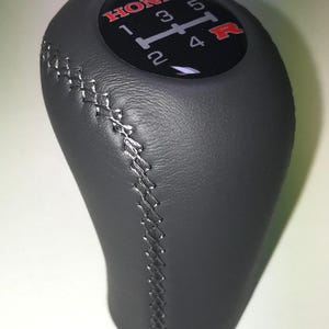 May include: A dark grey Honda gear stick knob with silver stitching. The top has a black face with the Honda logo in red, and white numbers indicating gear positions.