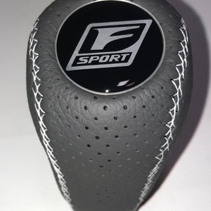 Peut inclure: Pommeau de levier de vitesse en cuir noir avec surpiqûres blanches et un logo F SPORT argenté.