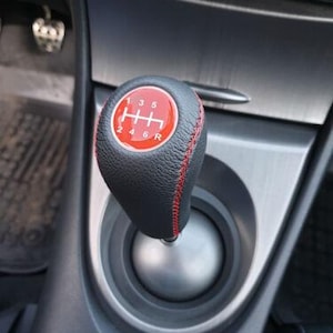 Op de afbeelding: Zwarte lederen versnellingspookknop met rode stiksels en een rood midden. Het midden toont een schakelpatroon met cijfers en letters. De knop is geplaatst in een zilveren en zwarte console.