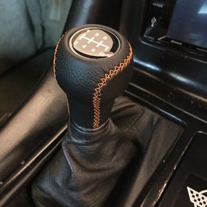 Könnte beinhalten: Ein schwarzer Leder-Schalthebelknauf mit orangefarbenen Nähten. Der Knauf hat ein silbernes Schaltmuster mit den Zahlen 1 bis 5 und R.