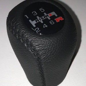 May include: Black leather gear shift knob with a black and silver Nismo logo and red R on a black background. The knob has a stitched seam and numbers 1 through 6.